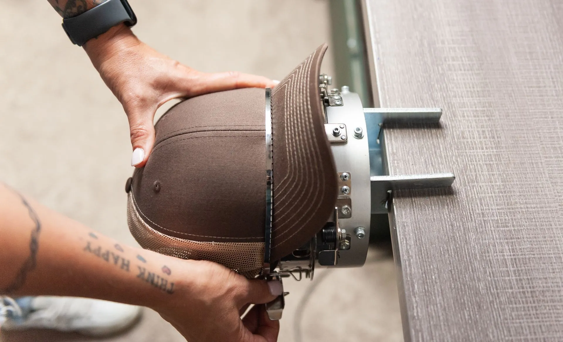 Person holding a brown cap in an embroidery hoop attached to a machine, preparing for customization. Tattoo and watch visible on wrist.