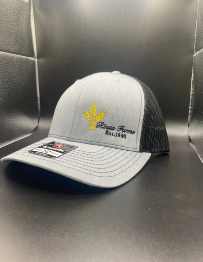 Gray and black baseball cap with a yellow wheat logo and "Rinax Farms Est. 1948" embroidered on the front. Cap is displayed on a black surface.