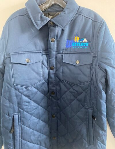 Blue quilted jacket with "Westlock Motors" logo on the chest, featuring snap buttons and flap pockets, displayed on a hanger against a light backdrop.