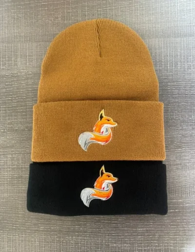 Two beanies on a textured surface; one brown and one black, each featuring an embroidered orange and white fox logo on the front.